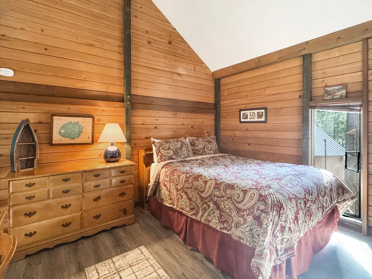 A warmly lit bedroom featuring a bed positioned near a window, a dresser, and a lamp. The window offers inviting views of the outdoors, suggesting a direct connection to nature and the activities just beyond the cabin.