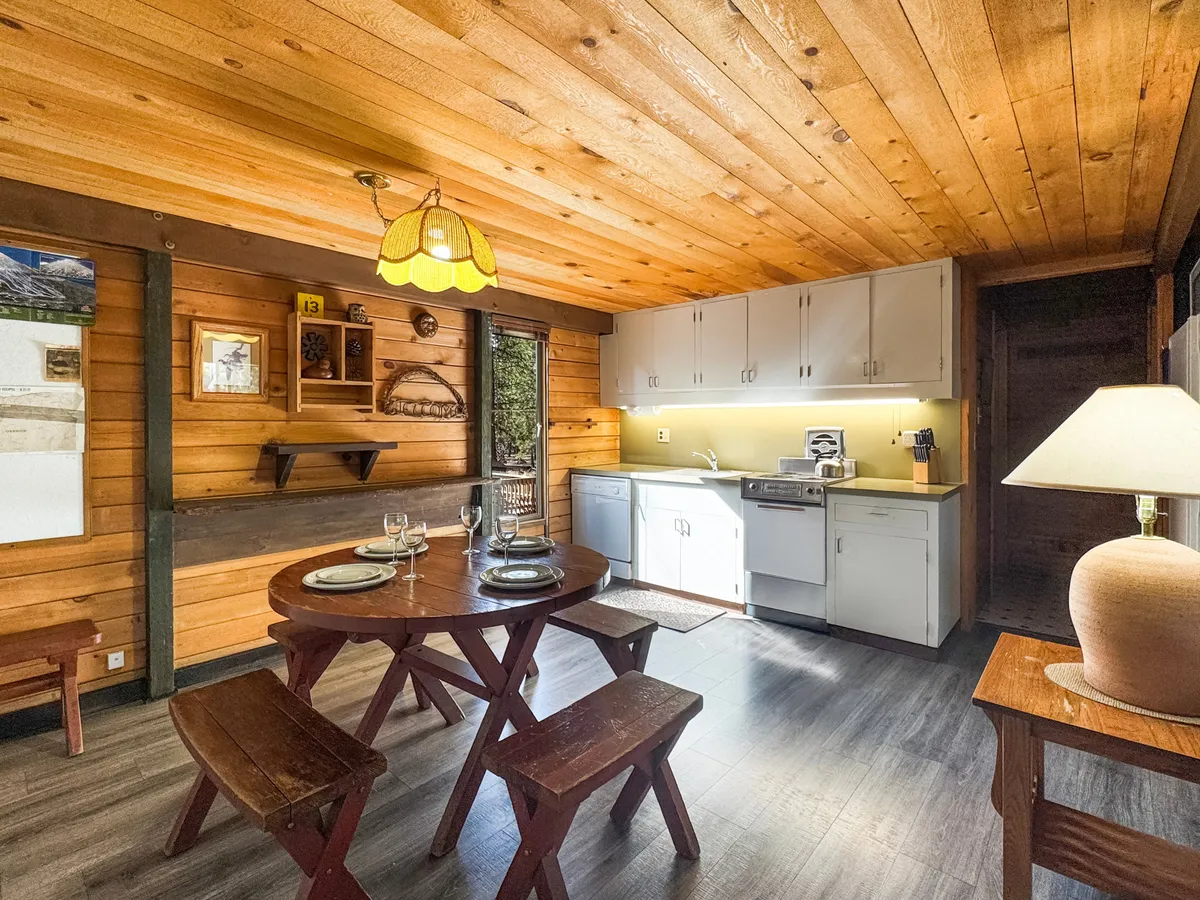 The image showcases a warm, inviting interior with wooden walls and an open layout connecting the dining area and kitchen. A wooden dining table sits under a cozy glass lamp, creating a welcoming space perfect for family meals and social gatherings. The overall ambiance is classic cabin comfort, designed for intimate moments and relaxed mornings with coffee.