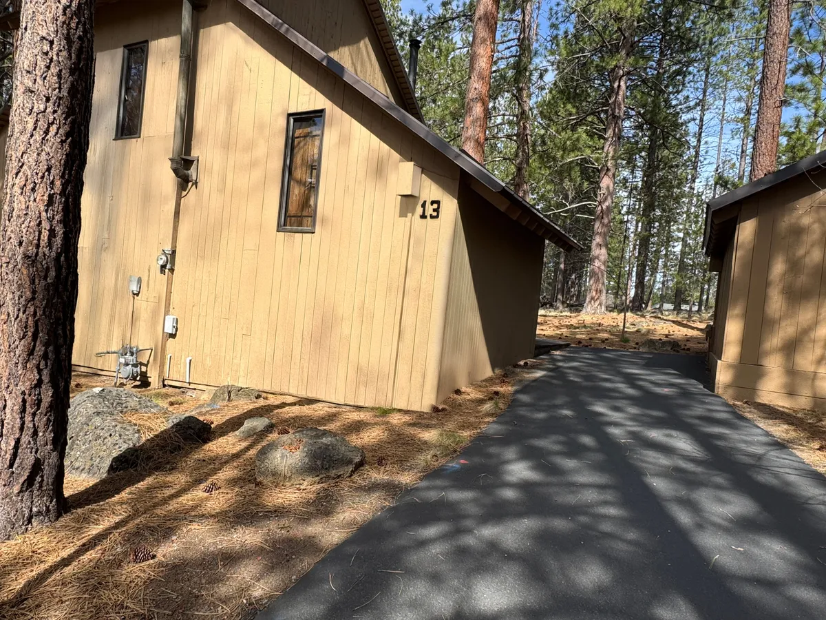 The front of the house numbered 13, with a tranquil pathway leading through pine trees directly to the entrance. The scene is set in inviting natural light, conveying an easy transition from home to the outdoors.