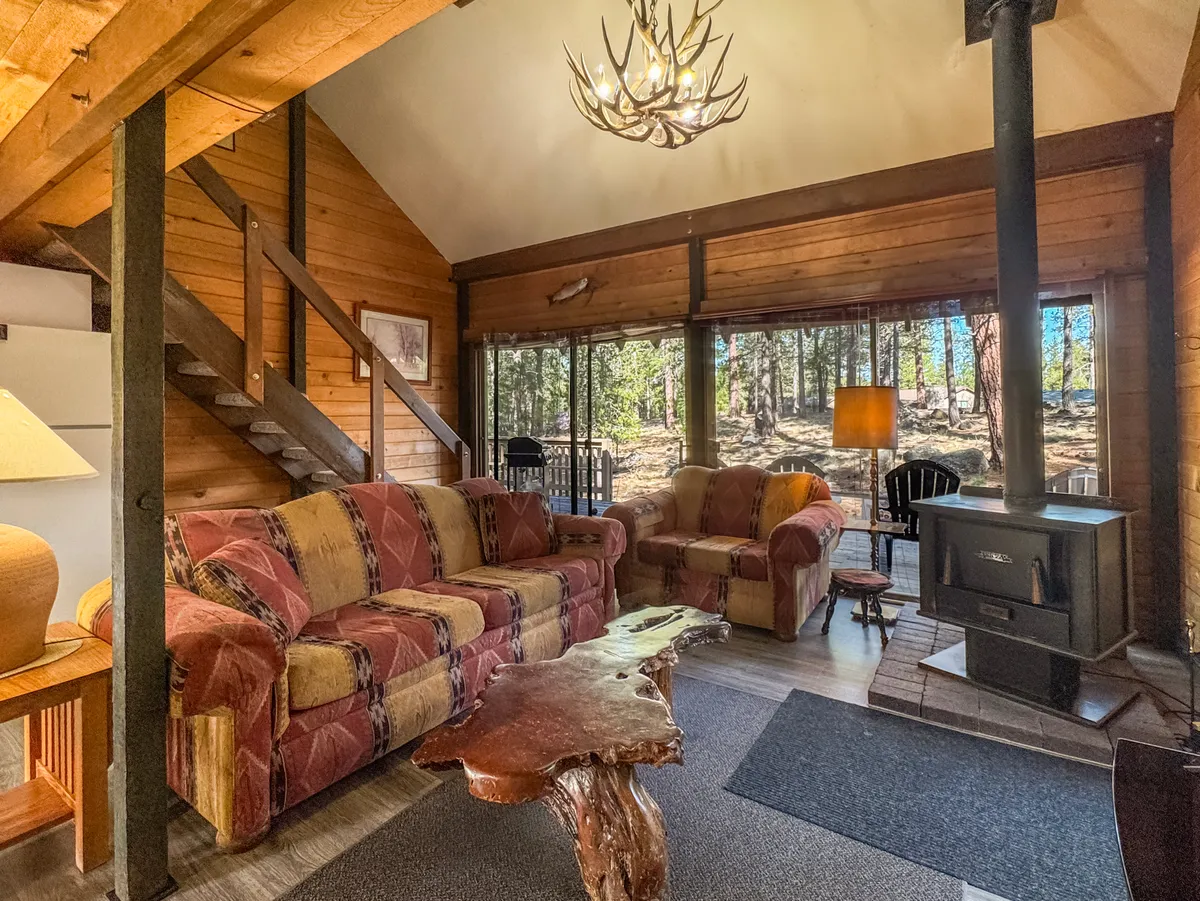This image captures the main living area of the cabin, featuring inviting wooden furniture, a prominent fireplace, large windows that fill the space with natural light, and a rustic chandelier overhead. The open arrangement encourages socializing and relaxation, creating a welcoming atmosphere for gathering with family and friends.