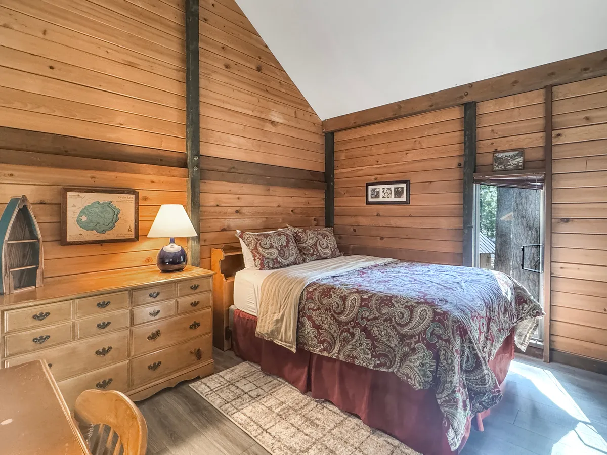 This image highlights a comfortable sleeping space with patterned bedding, a wooden dresser, and a bedside lamp. The presence of sunlight streaming in and the serene atmosphere suggest a restful retreat, which complements the description of the cabin's cozy sleeping options.