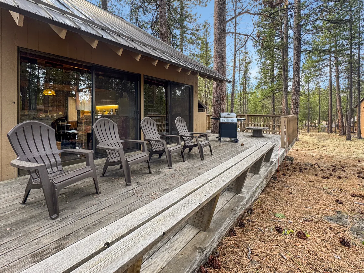 A cozy deck setup with Adirondack chairs and a grill, set amidst tall pine trees. The scene captures moments like socializing, barbecuing, or simply enjoying the outdoors in a relaxed, inviting atmosphere.