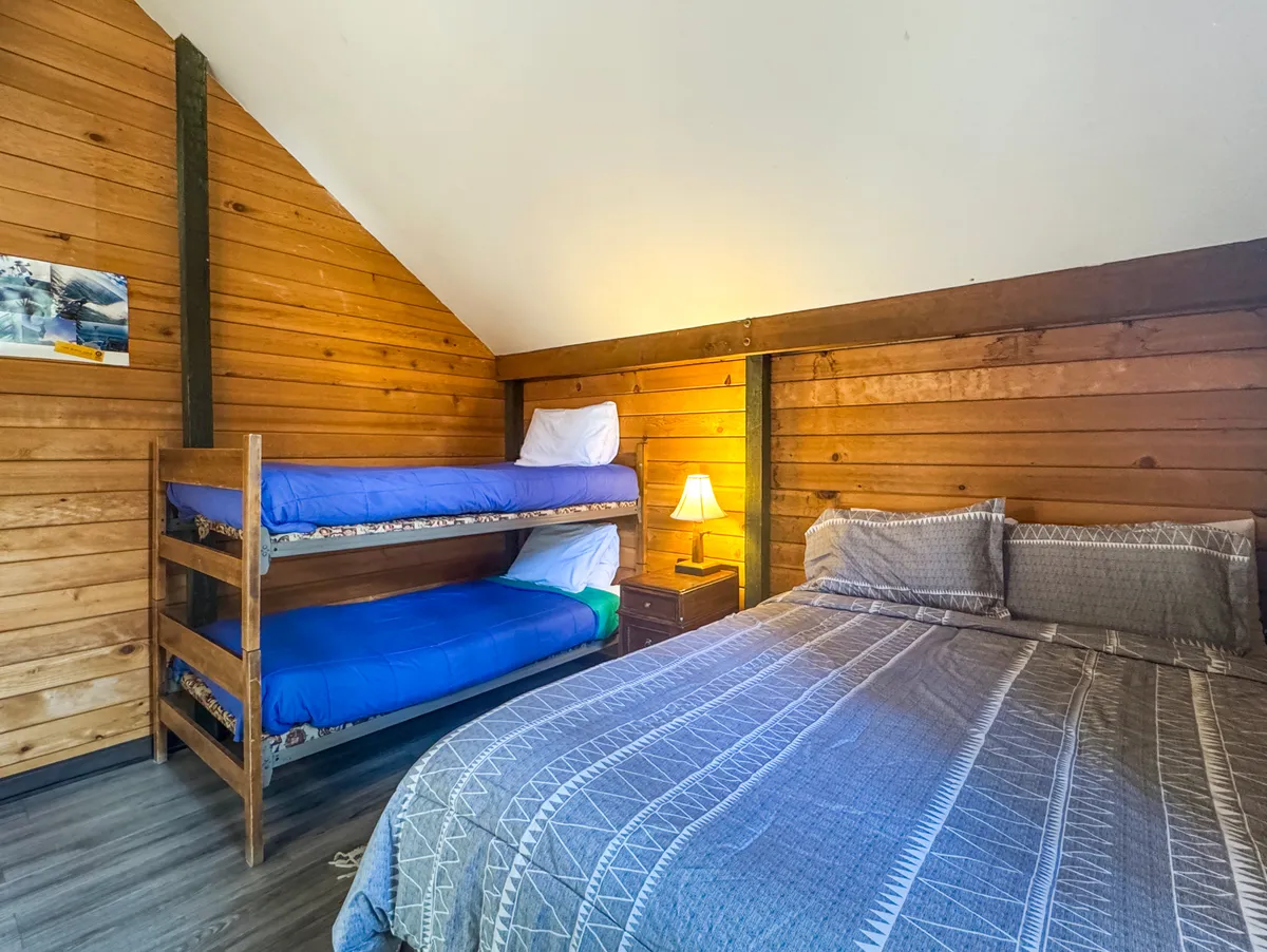 This image features a warm, inviting main bedroom, prominently showcasing a double bed and a set of bunk beds positioned beside a nightstand with a lamp. The room exudes a family-friendly atmosphere, ideal for group stays, and hints at playful, communal bonding with features like vintage posters and cozy bedding.