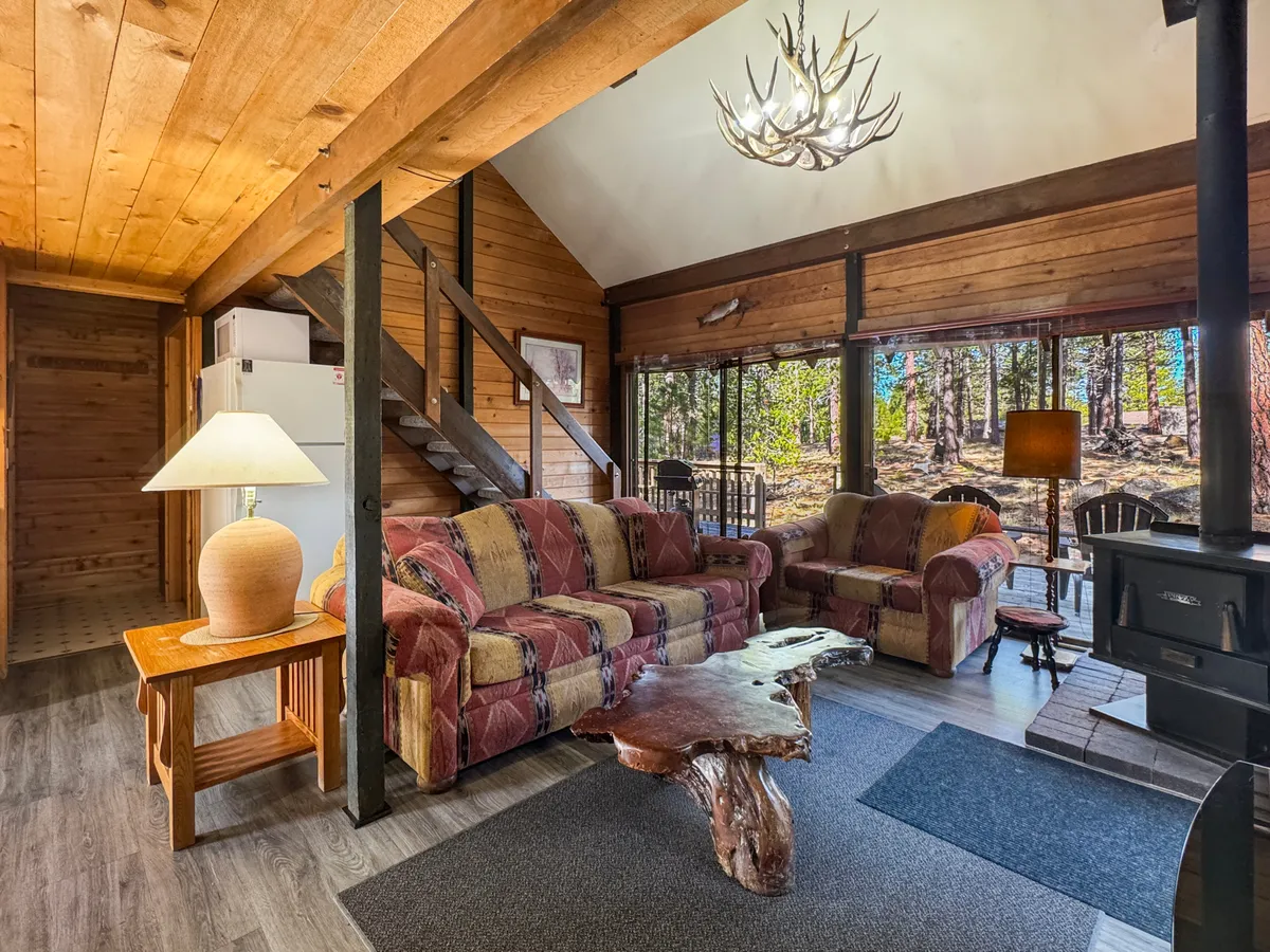 A warm, welcoming living area with comfortable sofas, wooden beams overhead, an antler chandelier, and a large coffee table. The space offers a relaxing spot to unwind after outdoor adventures, with large windows that hint at the natural setting just outside.