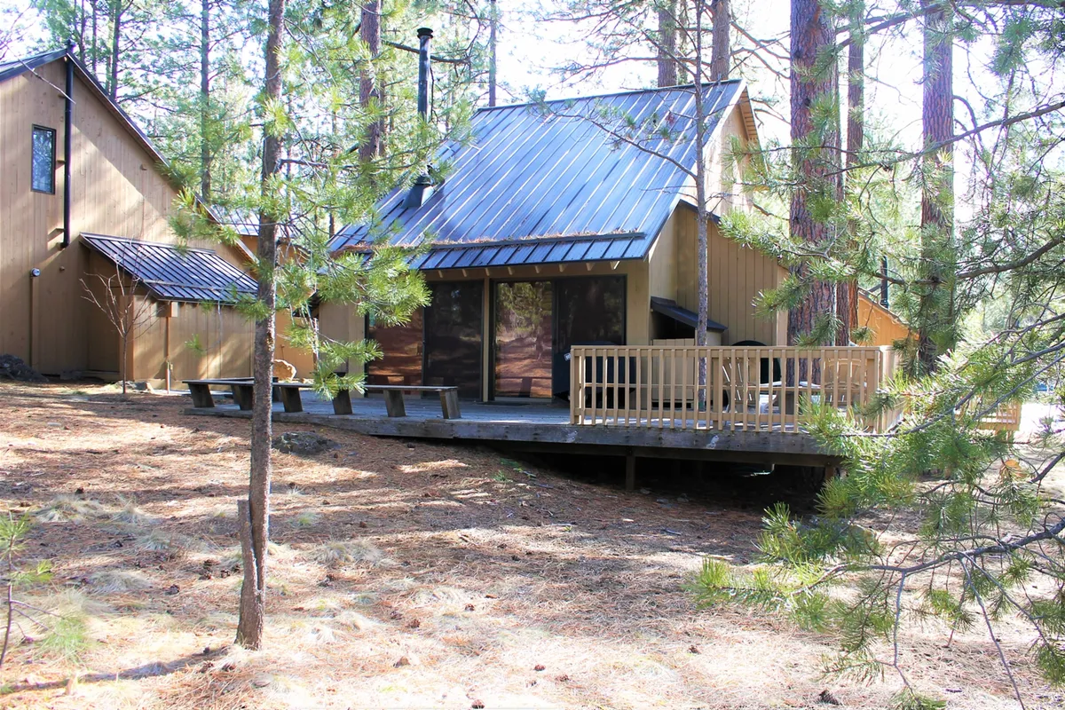 This image features a welcoming wooden deck surrounded by pine trees, with large windows and a sloped roof in view. The tranquil outdoor setting suggests easy access to nature and resort relaxation, all just steps from the cabin.